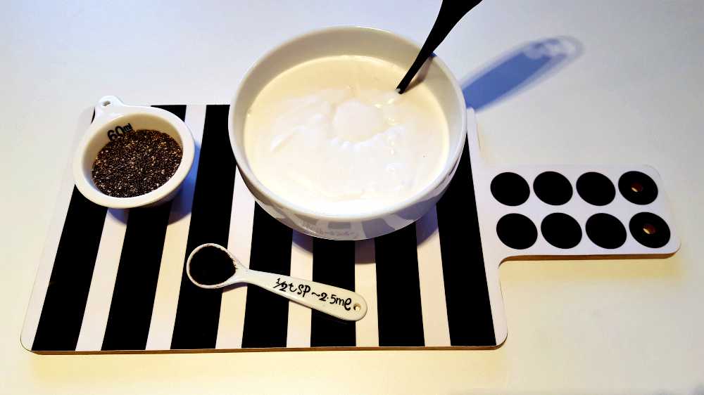 Skål med yoghurt och en sked på en randig skärbräda med chiafrön i en liten skål.